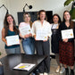 nail-technician-course-certificates-holding. Five women standing indoors and holding printed certificates. Two women wear aprons, and the setting appears to be a room with a black table, wall-mounted lamps, and a plant visible.