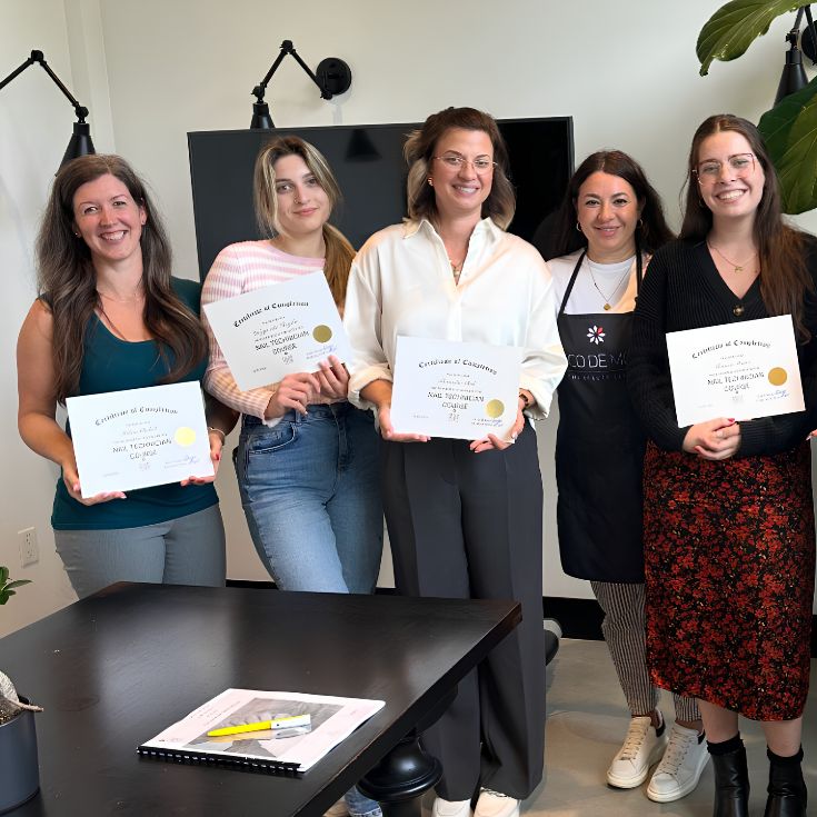 nail-technician-course-certificates-holding. Five women standing indoors and holding printed certificates. Two women wear aprons, and the setting appears to be a room with a black table, wall-mounted lamps, and a plant visible.